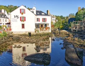 A charming riverside village scene featuring a white building with red window shutters and the sign 'Moulin du Grand Poulguin' on its wall. Surrounding the building, there is a small stream with large rocks and calm water that reflects the house. The area is lush with greenery, and other quaint buildings can be seen in the background, enhancing the picturesque view.