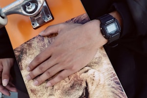 A hand with a black digital wristwatch rests on a skateboard. The skateboard features a lion image on its deck and has an orange color. The axel and wheel of the skateboard are visible, with a metallic silver finish.