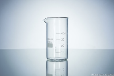 A clear glass beaker with measurement markings sits on a reflective surface. The background is a soft gradient from light grey to white, emphasizing the transparency and cleanliness of the beaker.