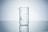 A clear glass beaker with measurement markings sits on a reflective surface. The background is a soft gradient from light grey to white, emphasizing the transparency and cleanliness of the beaker.
