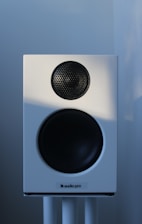 A white audio speaker with a black mesh-covered tweeter and a larger black subwoofer is mounted on a stand against a softly lit background.