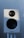 A white audio speaker with a black mesh-covered tweeter and a larger black subwoofer is mounted on a stand against a softly lit background.
