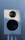 A white audio speaker with a black mesh-covered tweeter and a larger black subwoofer is mounted on a stand against a softly lit background.