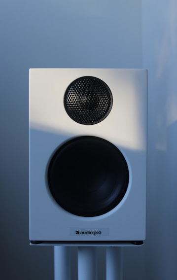 A white audio speaker with a black mesh-covered tweeter and a larger black subwoofer is mounted on a stand against a softly lit background.