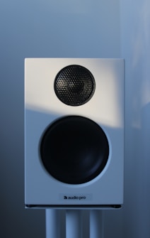 A white audio speaker with a black mesh-covered tweeter and a larger black subwoofer is mounted on a stand against a softly lit background.