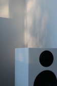 A modern white speaker with two visible black speaker grilles, set against a background that is softly blurred. Natural light casts gentle shadows and highlights on the wall and speaker surface.