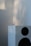 A modern white speaker with two visible black speaker grilles, set against a background that is softly blurred. Natural light casts gentle shadows and highlights on the wall and speaker surface.