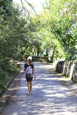 A person walks leisurely down a sun-dappled path surrounded by lush green trees. They are wearing a backpack and a cap, suggesting a casual outing or hike. The pathway is flanked by stone walls, enhancing the serene natural setting.