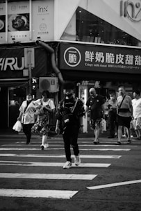 A group of people walking across a pedestrian crossing in an urban street setting. Some individuals are carrying shopping bags. Buildings are visible in the background with various signs, some with Asian characters and one advertising crispy milk donuts. The scene is in black and white, emphasizing contrasts and shadows.