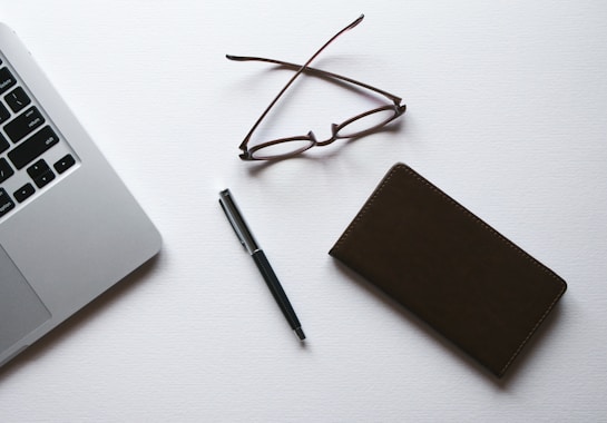 A minimalist arrangement featuring the corner of a laptop, a pair of eyeglasses, a pen, and a closed notebook placed on a white surface.