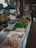 A market stall is filled with a variety of fresh produce and goods. There are trays of eggs, garlic, and an assortment of vegetables such as bell peppers, green onions, carrots, and leafy greens. In the background, people are walking around and browsing, with some stall keepers assisting customers.