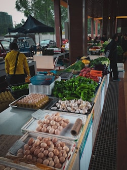 A market stall is filled with a variety of fresh produce and goods. There are trays of eggs, garlic, and an assortment of vegetables such as bell peppers, green onions, carrots, and leafy greens. In the background, people are walking around and browsing, with some stall keepers assisting customers.