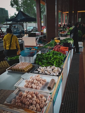 A market stall is filled with a variety of fresh produce and goods. There are trays of eggs, garlic, and an assortment of vegetables such as bell peppers, green onions, carrots, and leafy greens. In the background, people are walking around and browsing, with some stall keepers assisting customers.
