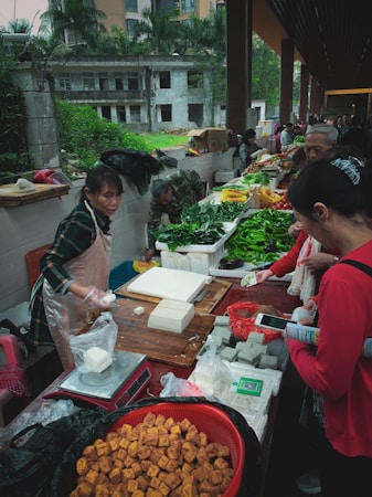 A busy outdoor market scene with various people interacting around a stall selling fresh produce and tofu. The table is filled with green vegetables, blocks of white and gray tofu, and a basket of fried tofu cubes. A woman in an apron is weighing products on a scale while customers gather around, some holding shopping bags and mobile phones. The background shows greenery and buildings under a covered area.