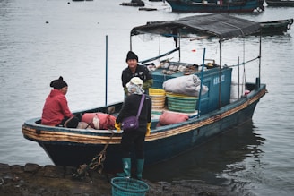 A small fishing boat is docked at the edge of a body of water. Three people are on the boat, dressed in warm clothing. Various sacks and baskets with goods are scattered around the deck. The scene suggests a fishing community setting with overcast weather.