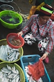 A person wearing a colorful beanie and plaid shirt is selecting seafood at an outdoor market. Various containers filled with fresh fish and other sea creatures are displayed. The ground is wet, suggesting it's a fresh and lively environment. The person is holding a bag and appears to be in the process of purchasing or sorting through the seafood.