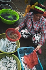 A person wearing a colorful beanie and plaid shirt is selecting seafood at an outdoor market. Various containers filled with fresh fish and other sea creatures are displayed. The ground is wet, suggesting it's a fresh and lively environment. The person is holding a bag and appears to be in the process of purchasing or sorting through the seafood.