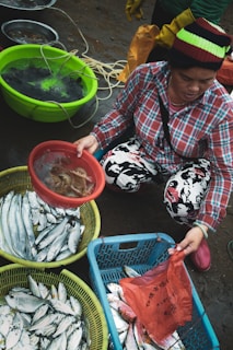 A person wearing a colorful beanie and plaid shirt is selecting seafood at an outdoor market. Various containers filled with fresh fish and other sea creatures are displayed. The ground is wet, suggesting it's a fresh and lively environment. The person is holding a bag and appears to be in the process of purchasing or sorting through the seafood.