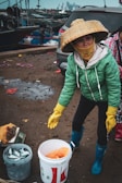 A person wearing a green jacket and a large, woven hat is standing outside near a car and some fishing equipment. The person is holding a pair of yellow gloves and is surrounded by buckets, one of which contains fish. The background shows a dock with several boats and industrial structures in the distance.