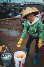 A person wearing a green jacket and a large, woven hat is standing outside near a car and some fishing equipment. The person is holding a pair of yellow gloves and is surrounded by buckets, one of which contains fish. The background shows a dock with several boats and industrial structures in the distance.