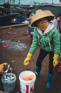 A person wearing a green jacket and a large, woven hat is standing outside near a car and some fishing equipment. The person is holding a pair of yellow gloves and is surrounded by buckets, one of which contains fish. The background shows a dock with several boats and industrial structures in the distance.