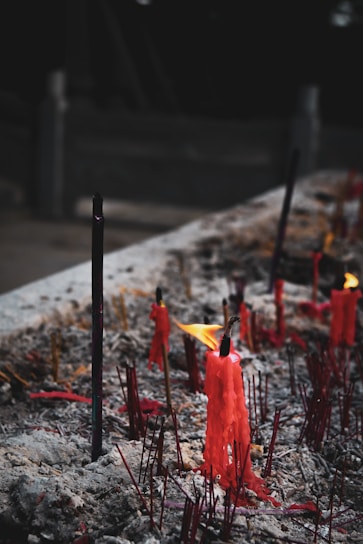 Red candles, some lit with small flames, are surrounded by ashes and remnants of burned incense sticks against a dark background. The scene appears intimate and contemplative, with melted wax forming abstract shapes.