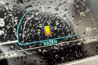 A digital car dashboard displaying a battery icon with a highlight on regenerative braking, surrounded by raindrops on the glass, indicating a rainy weather setting.
