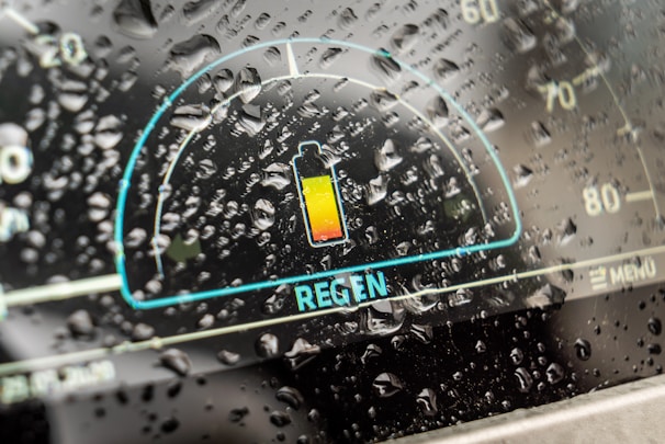A digital car dashboard displaying a battery icon with a highlight on regenerative braking, surrounded by raindrops on the glass, indicating a rainy weather setting.