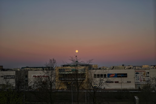 A shopping mall is visible in the foreground, with various store signs such as Media Markt, KFC, and others. In the background, there is a clear sky with a gradient from blue to pink, and a full moon is centrally positioned above the mall. Leafless trees are silhouetted against the sky, adding to the composition.