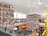 A large, modern library with towering bookshelves filled with colorful books. The room has a spacious, open layout with an escalator on the left and several people walking around or standing. The ceiling is high with an artistic light fixture composed of spherical lights. There are large windows allowing natural light to illuminate the space.