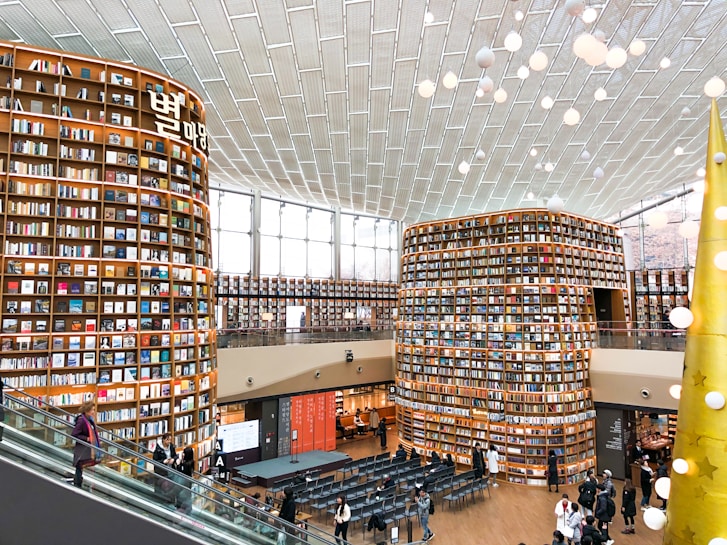 A large, modern library with towering bookshelves filled with colorful books. The room has a spacious, open layout with an escalator on the left and several people walking around or standing. The ceiling is high with an artistic light fixture composed of spherical lights. There are large windows allowing natural light to illuminate the space.