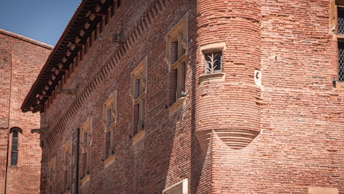 A historic brick building with intricate masonry featuring a prominent circular turret. The facade includes several tall, narrow windows with shutters. The sun casts shadows, highlighting the texture of the bricks.