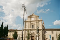 A historic church facade with intricate carvings and a prominent statue in front. The sky is partially cloudy, suggesting a bright and pleasant day. There are a few trees and buildings surrounding the church, along with a tall, ornate streetlamp.