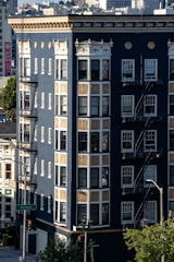 A multi-story urban building with a dark blue facade and ornate gold and white detailing. The windows are numerous and framed in white, and there are fire escape staircases on the side. In the foreground, a street sign indicates Fulton Street. The background shows a densely packed cityscape.