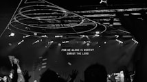 A black and white image capturing a live concert or worship event with people raising their hands. Stage lights and circular hanging structures are visible above the crowd. Text displayed on a large screen reads 'FOR HE ALONE IS WORTHY CHRIST THE LORD'.