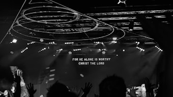 A black and white image capturing a live concert or worship event with people raising their hands. Stage lights and circular hanging structures are visible above the crowd. Text displayed on a large screen reads 'FOR HE ALONE IS WORTHY CHRIST THE LORD'.