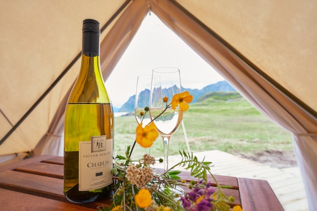 A bottle of Chablis wine is placed on a wooden table inside a tent. Beside it are two wine glasses, one filled with wine and both decorated with yellow and white flowers. The tent is pitched on a grassy field with mountains visible in the background.