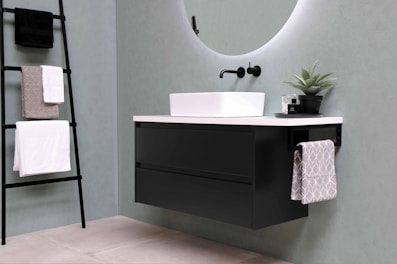 A modern bathroom setup featuring a sleek, rectangular sink mounted on a black floating cabinet with clean lines. A round, backlit mirror is mounted above the sink. A matte black wall-mounted faucet adds to the contemporary design. A ladder-style towel rack holds several neatly folded towels, while a small potted plant sits on the countertop for a touch of greenery.