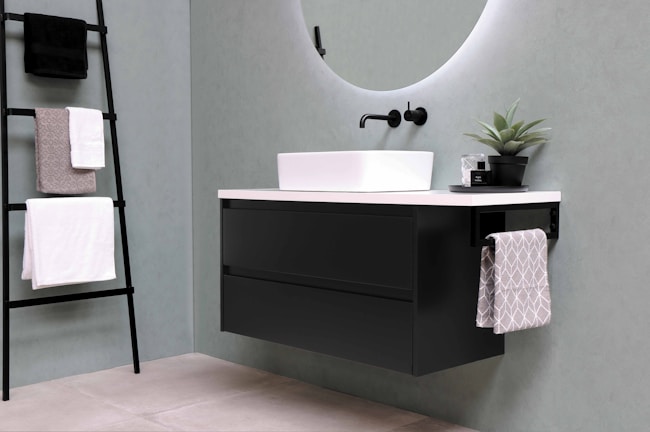 A modern bathroom setup featuring a sleek, rectangular sink mounted on a black floating cabinet with clean lines. A round, backlit mirror is mounted above the sink. A matte black wall-mounted faucet adds to the contemporary design. A ladder-style towel rack holds several neatly folded towels, while a small potted plant sits on the countertop for a touch of greenery.