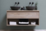 A modern bathroom vanity with two black stone vessel sinks and wall-mounted faucets above them. Between the sinks, there is a small bottle of soap and toothbrushes in a holder. Underneath, an open shelf holds neatly folded white and black towels and a wire basket with additional folded towels. A small green plant in a transparent vase is placed to the right of the sink.