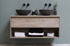 A modern bathroom vanity with two black stone vessel sinks and wall-mounted faucets above them. Between the sinks, there is a small bottle of soap and toothbrushes in a holder. Underneath, an open shelf holds neatly folded white and black towels and a wire basket with additional folded towels. A small green plant in a transparent vase is placed to the right of the sink.