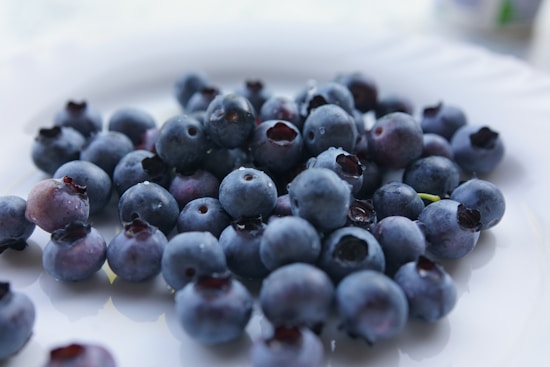 A pile of fresh blueberries sits on a clean, white surface, with varying shades of deep blue and purple. The berries are grouped closely together, showcasing their smooth, round shapes and natural variation in size and color. A few berries display slight imperfections or natural dimples.