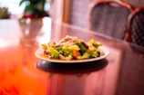 A plate of stir-fried vegetables, including broccoli, carrots, and mushrooms, sits on a reflective glass table. The background features blurred dining chairs and plants, creating a warm, inviting atmosphere.