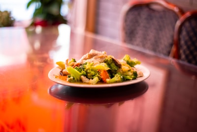 A plate of stir-fried vegetables, including broccoli, carrots, and mushrooms, sits on a reflective glass table. The background features blurred dining chairs and plants, creating a warm, inviting atmosphere.
