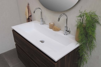 A modern bathroom sink setup with two chrome faucets, a white rectangular basin, and a dark wooden cabinet underneath. Towels hang on the wall to the left, and a potted green plant is on the right. A bathroom mirror is partially visible above the sink, and small bathroom accessories, including a sponge and container, are placed on the sink's countertop.