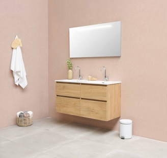 A minimalist bathroom with a floating wooden vanity featuring two sinks. Above the sinks is a wide, rectangular mirror. A small potted plant and soap dispenser are placed on the counter. On the left wall, a white towel hangs beside a natural woven basket containing folded towels. The walls are a soft beige color, and the floor has large, light gray tiles. A small white trash bin is situated next to the vanity.