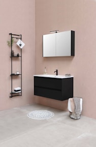 The bathroom features a modern, minimalist design with a black floating vanity and a white countertop, accompanied by a black faucet. Above the vanity, a wall-mounted mirror cabinet offers storage. There's a black metal shelf unit to the left, holding some decorative items, including folded towels and small plants. A wire basket containing towels is placed on the right side, while a round grey rug is positioned on the floor in front of the vanity. The color palette includes soft beige walls and light grey tiled flooring.