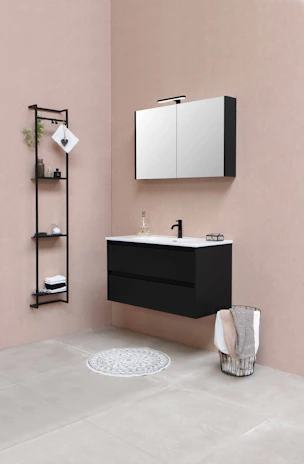 The bathroom features a modern, minimalist design with a black floating vanity and a white countertop, accompanied by a black faucet. Above the vanity, a wall-mounted mirror cabinet offers storage. There's a black metal shelf unit to the left, holding some decorative items, including folded towels and small plants. A wire basket containing towels is placed on the right side, while a round grey rug is positioned on the floor in front of the vanity. The color palette includes soft beige walls and light grey tiled flooring.