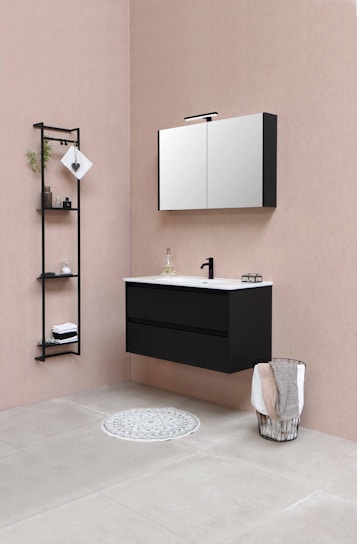 The bathroom features a modern, minimalist design with a black floating vanity and a white countertop, accompanied by a black faucet. Above the vanity, a wall-mounted mirror cabinet offers storage. There's a black metal shelf unit to the left, holding some decorative items, including folded towels and small plants. A wire basket containing towels is placed on the right side, while a round grey rug is positioned on the floor in front of the vanity. The color palette includes soft beige walls and light grey tiled flooring.