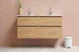 A modern bathroom setting featuring a wooden floating vanity with two chrome faucets. On the left, a tall, beige vase contains green foliage, while a small decorative object sits between the sinks. The background wall is painted in a light, warm tone, and a white, round pedal bin is placed on the floor to the right.